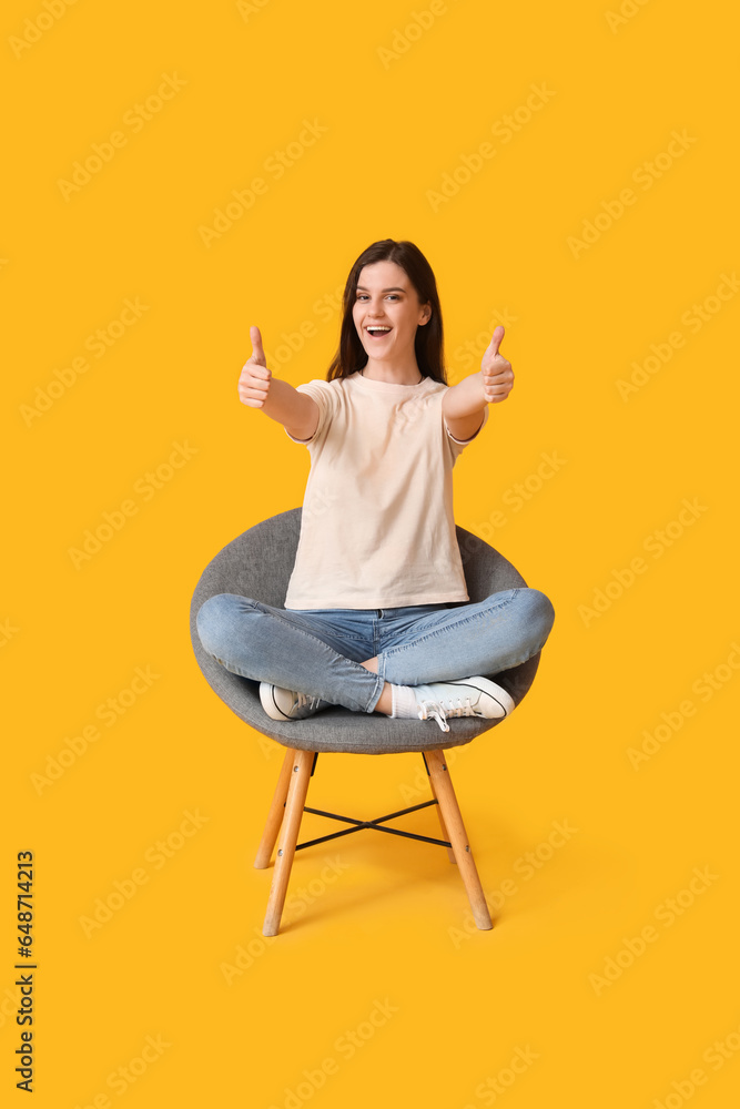 Young woman showing thumbs-up in armchair on yellow background