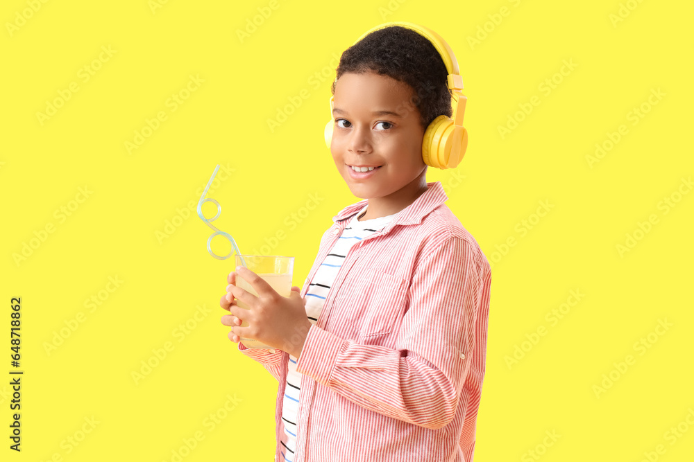 Little African-American boy in headphones with cup of fresh citrus juice on yellow background