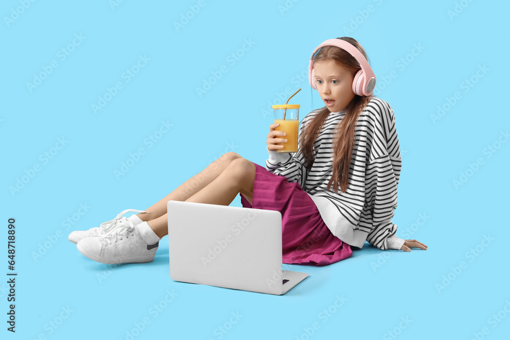 Little girl with glass of fresh citrus juice and laptop sitting on blue background