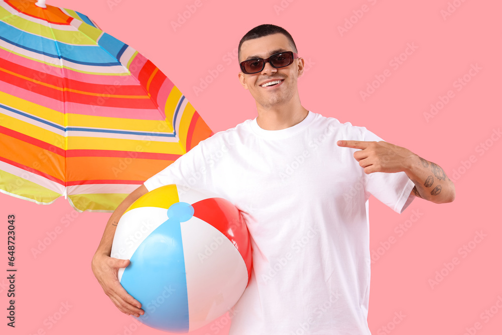 Happy young man with umbrella pointing at inflatable ball on pink background