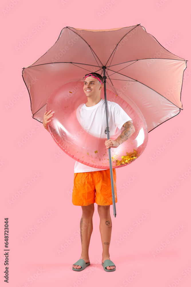 Happy young man with inflatable ring and umbrella on pink background