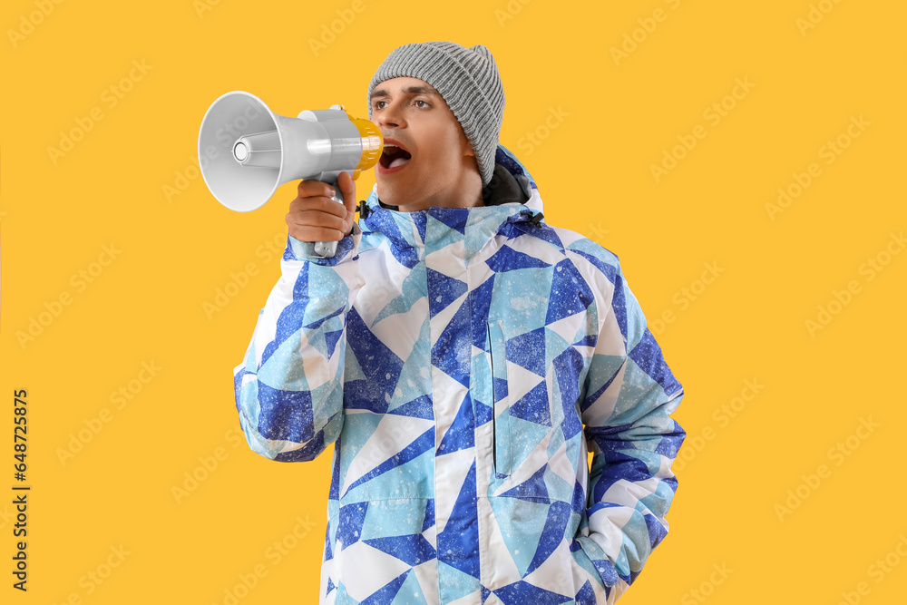 Young man in winter clothes shouting into megaphone on yellow background