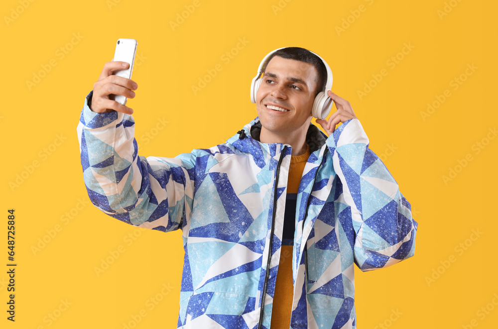 Young man in warm jacket with headphones taking selfie on yellow background