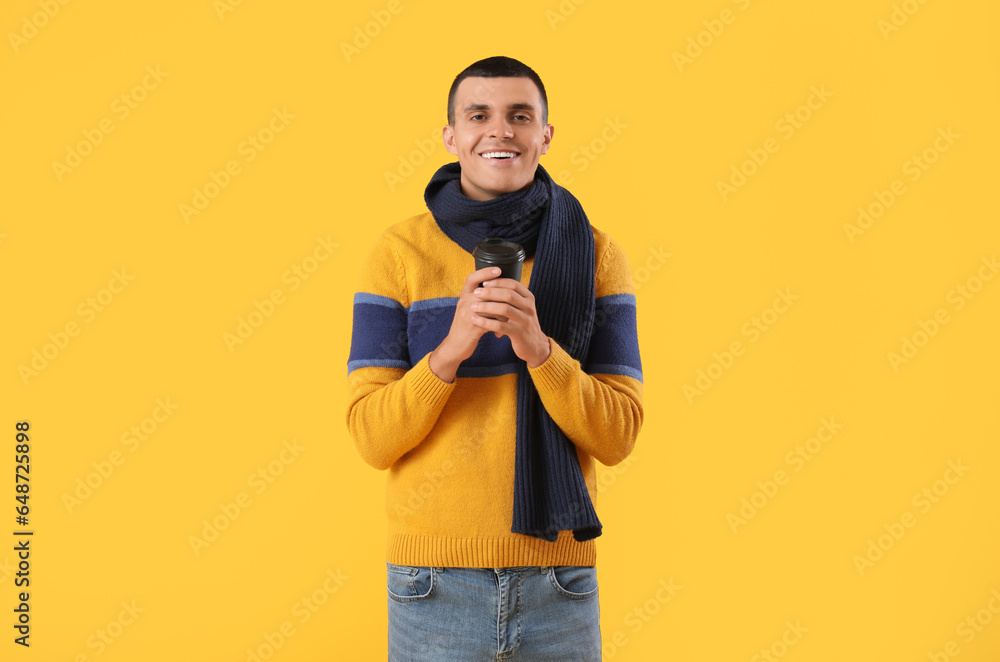 Young man in winter clothes with cup of coffee on yellow background