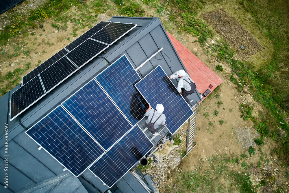 Technicians installing solar panel system on roof of house. Aerial view of men workers in helmets carrying photovoltaic solar module outdoors. Concept of alternative and renewable energy.