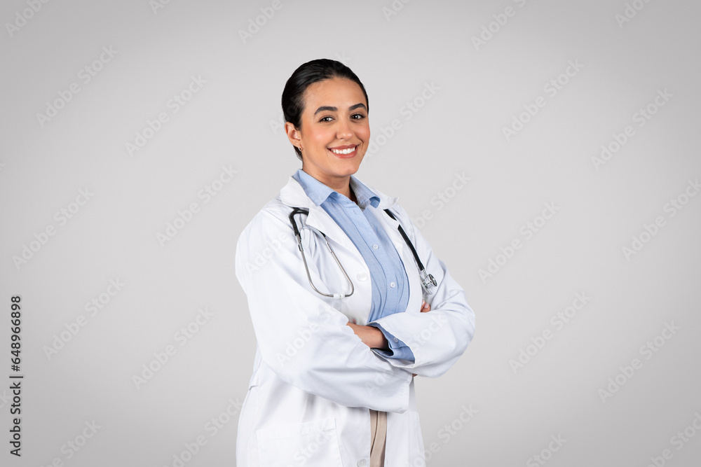 Medical worker. Cheerful brazilian doctor woman in white uniform smiling at camera while posing over grey background