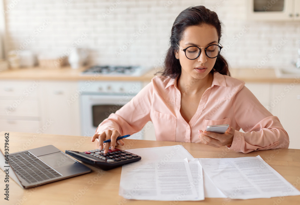 Woman using calculator and cellphone, counting bills or monthly expenses, sitting at table in kitchen