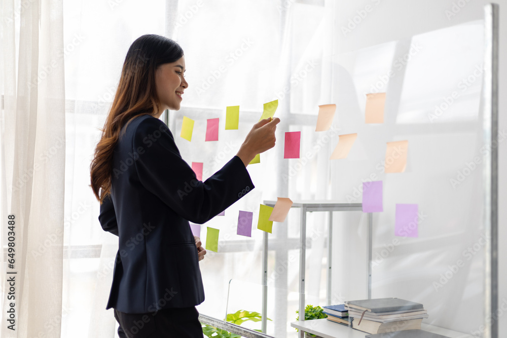 Asian businesswoman scheduling work strategy, planning work in the office with paper stickers on the glass.