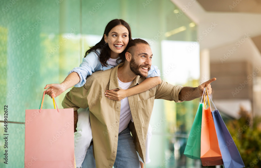 Glad young european guy with bags holds on back lady, point finger at copy space, have fun