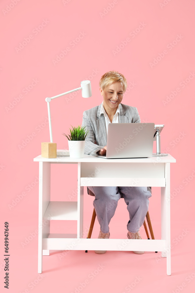 Mature female programmer working with laptop at table on pink background