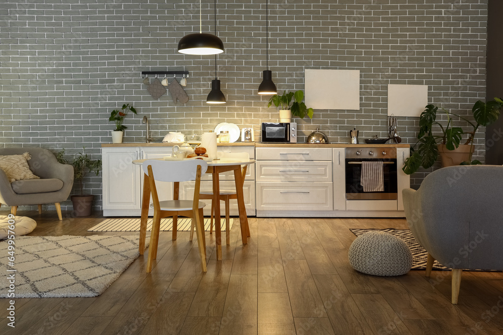 Interior of modern kitchen with glowing lamps, dining table, white counters and grey sofa at evening