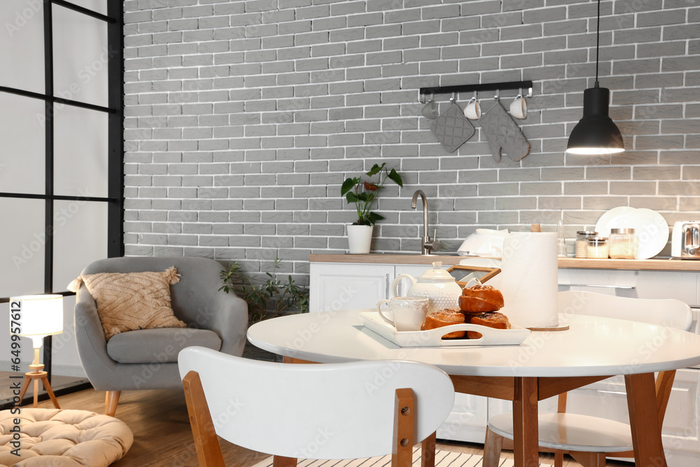 Interior of modern kitchen with glowing lamps, dining table, white counters and grey armchair at evening