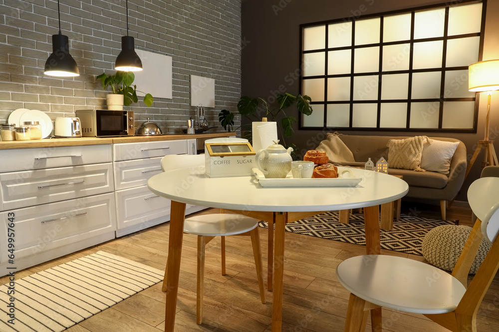 Interior of modern kitchen with glowing lamps, dining table, white counters and grey sofa at evening