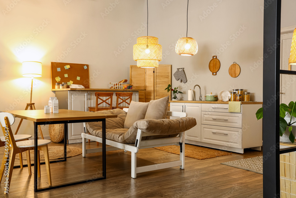 Interior of modern kitchen with glowing lamps, dining table, sofa and white counters at evening