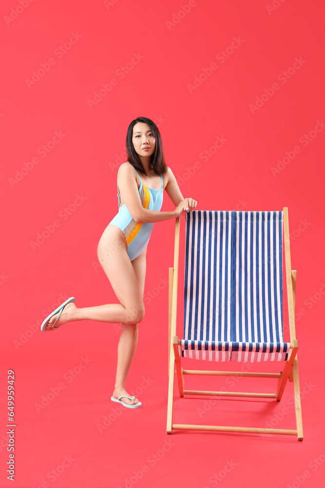 Beautiful Asian woman in swimsuit with deck chair on red background