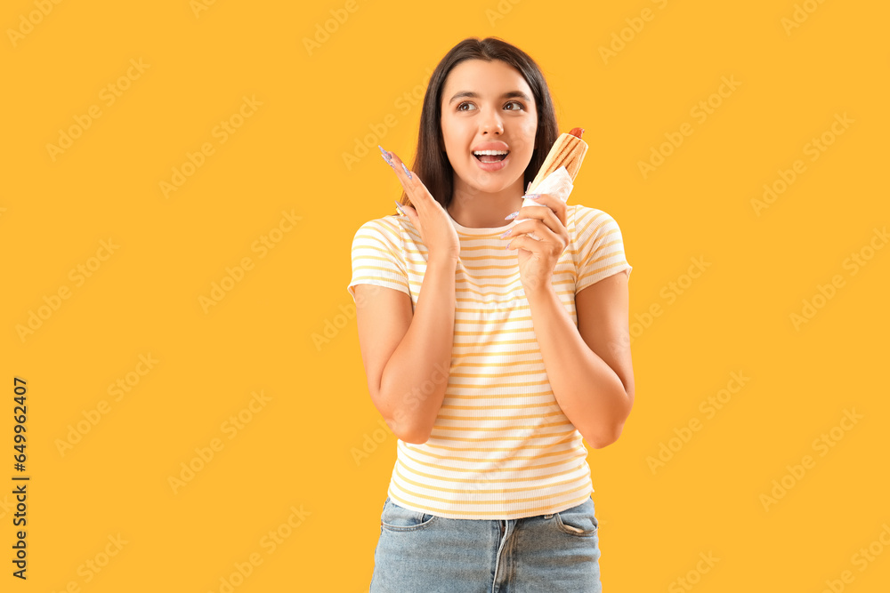 Happy young woman with tasty french hot dog on yellow background