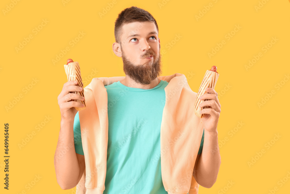 Portrait of doubtful young man with tasty hot dogs on yellow background