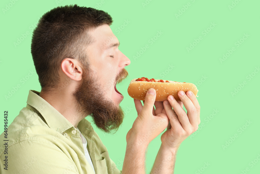 Portrait of handsome young man eating tasty hot dog on green background