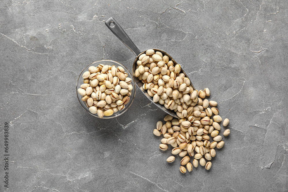 Glass bowl and metal scoop with tasty pistachio nuts on grey background