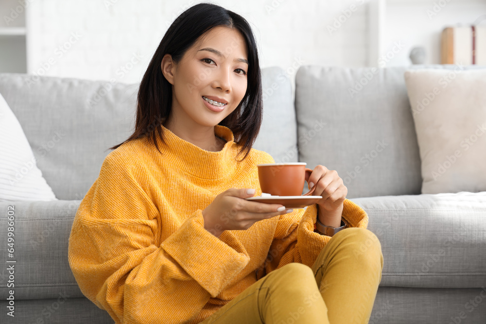 Beautiful young Asian woman with cup of tea in living room at home