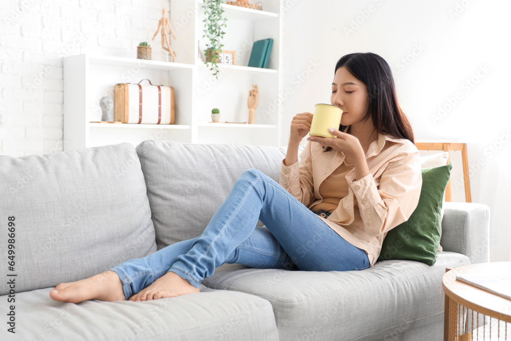 Beautiful young Asian woman with cup of tea sitting on sofa in living room