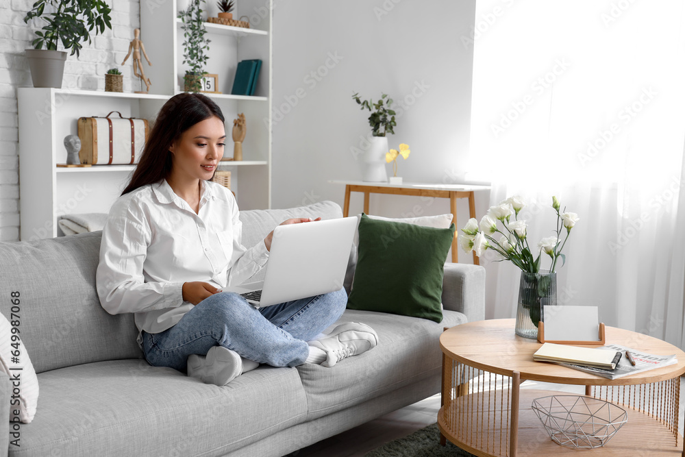 Beautiful young Asian woman with laptop sitting on sofa in living room