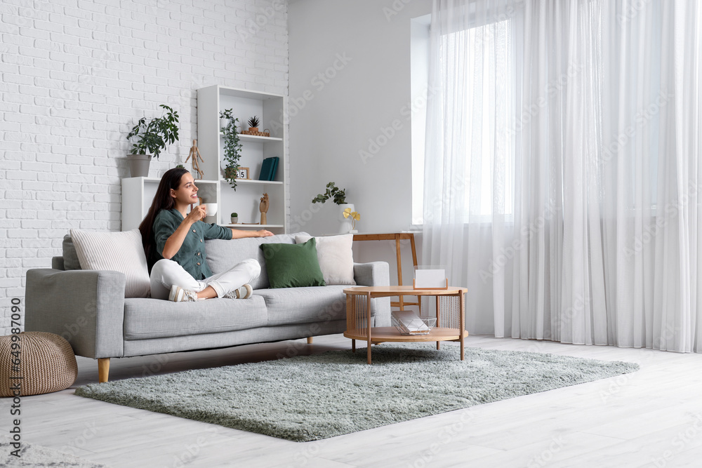 Beautiful young woman with cup of coffee sitting on sofa in living room