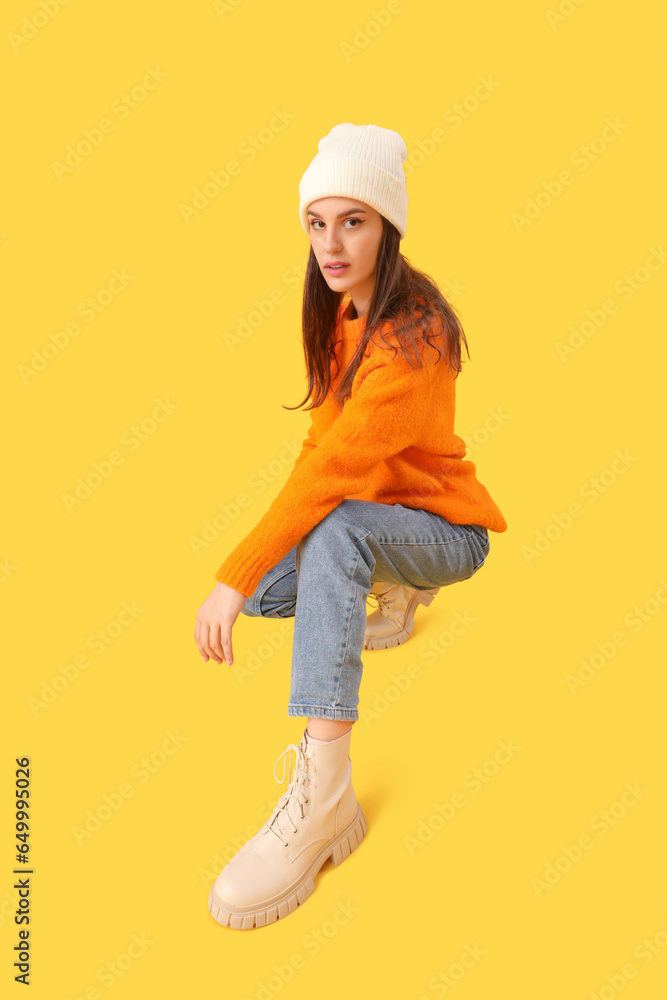 Young woman in winter clothes sitting on yellow background