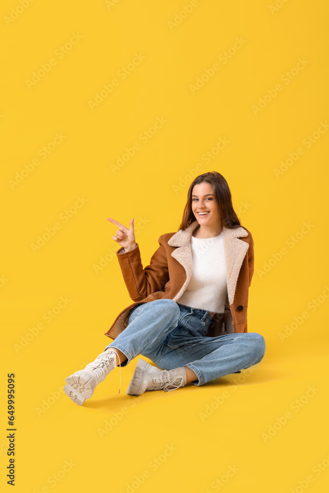 Young woman in winter clothes pointing at something on yellow background