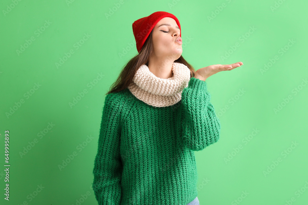 Young woman in winter clothes blowing kiss on green background