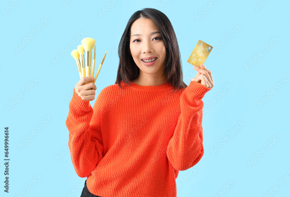 Young Asian woman with credit card and makeup brushes on blue background