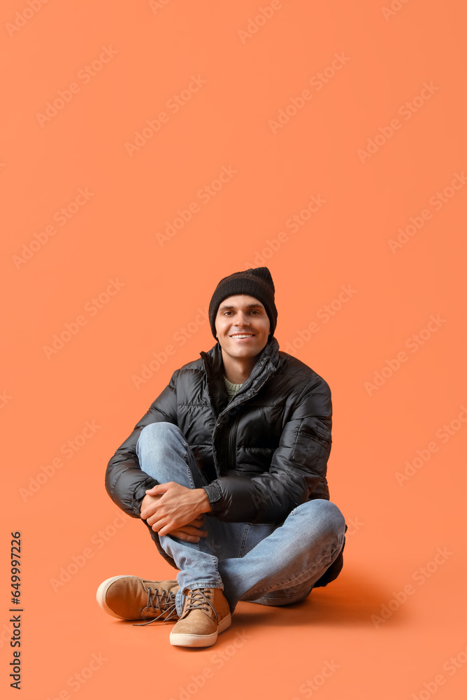 Young man in winter clothes sitting on orange background