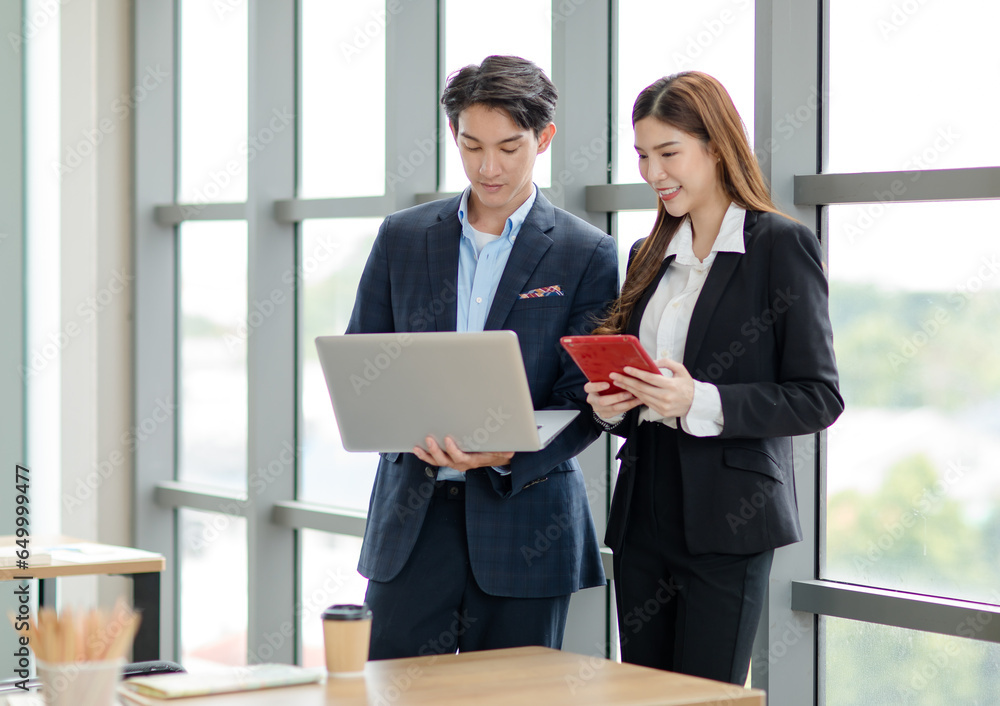 Asian professional successful female businesswoman and male businessman colleague employee in formal suit standing smiling together browsing surfing internet online via laptop and tablet in office