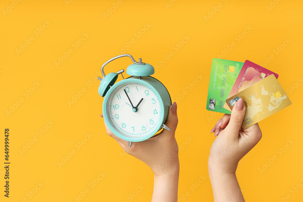 Female hands holding alarm clock and credit cards on yellow background