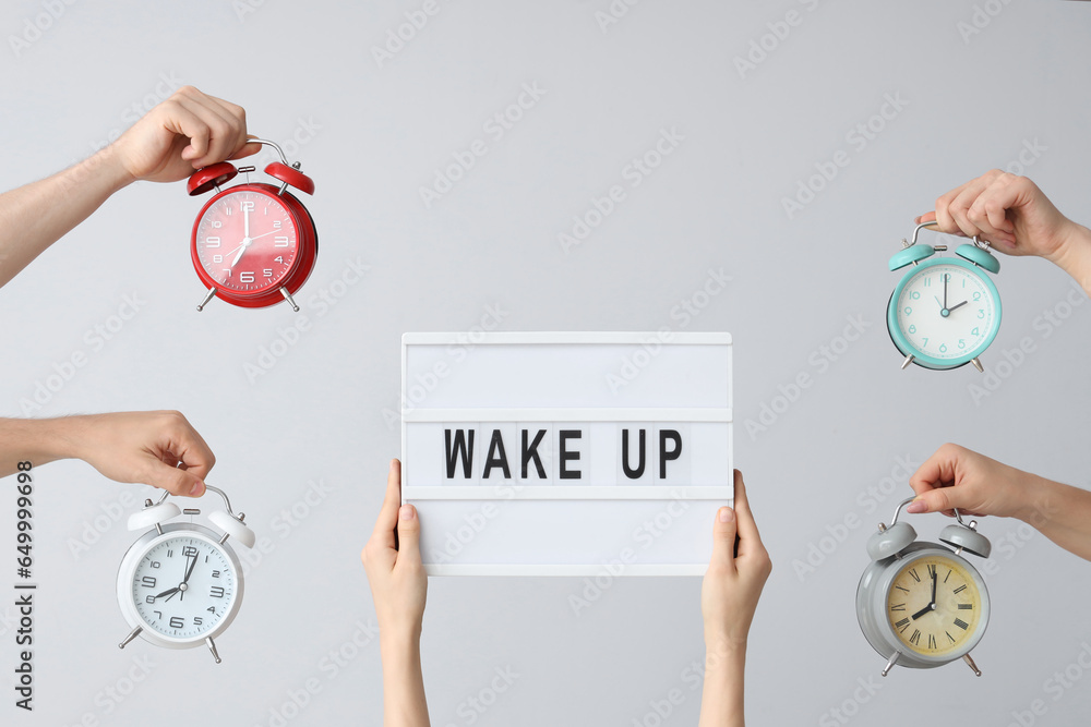 Hands holding different alarm clocks and board with text WAKE UP on grey background