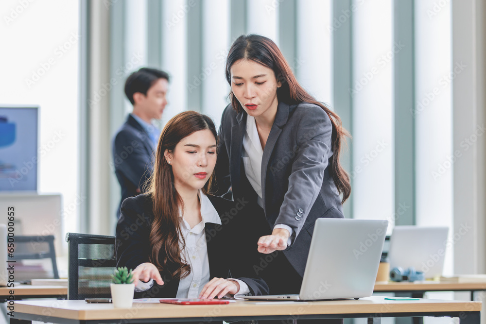 Asian professional successful female businesswoman supervisor mentor in formal suit standing smiling pointing help advising new colleague working with paperwork document at working station in office