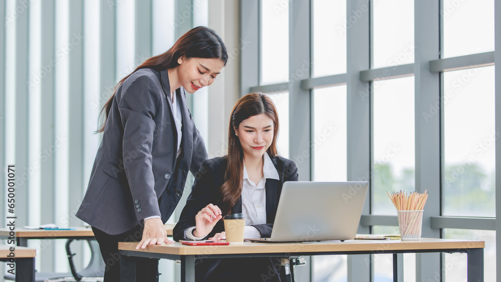 Asian professional successful female businesswoman supervisor mentor in formal suit standing smiling pointing help advising new colleague working with paperwork document at working station in office