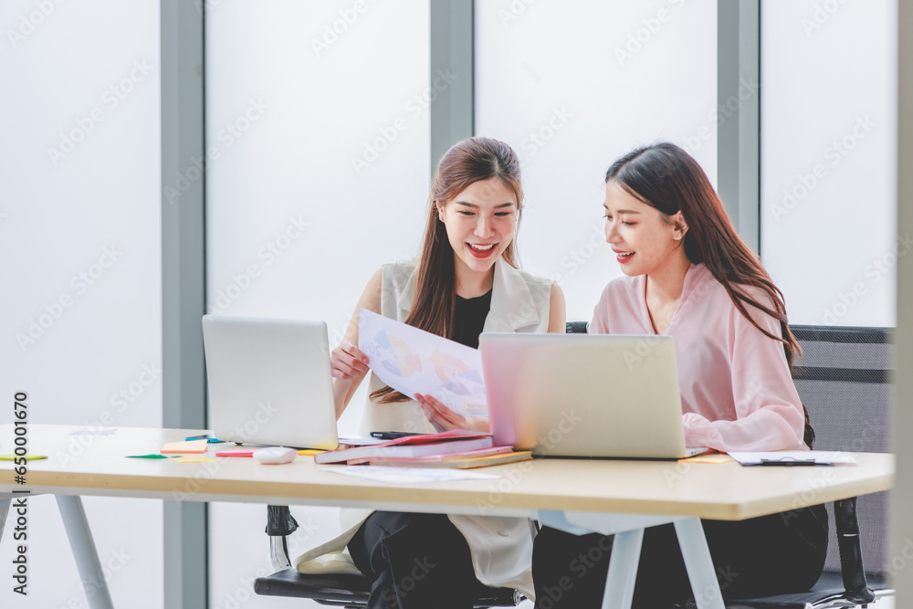 Two Asian professional successful young female businesswomen employee in casual outfit sitting working together with graph chart paperwork document report on workstation desk with laptop in office