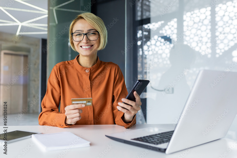 Portrait of young beautiful woman at workplace, businesswoman holding phone and bank credit card in hands, smiling and looking into camera, female employee booking online services and buying internet