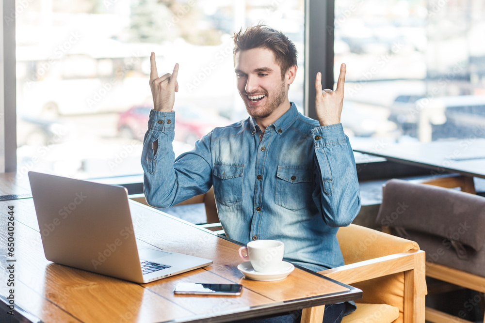 Portrait of crazy excited cool young man freelancer in blue jeans shirt working on laptop, showing rock and roll gesture to computer screen. Indoor shot near big window, cafe background.