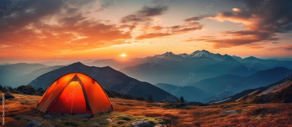 Vibrant orange tent beneath dramatic sunset in mountainous summer panorama