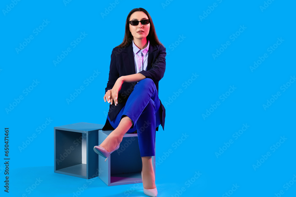 Young woman in stylish suit sitting on blue background