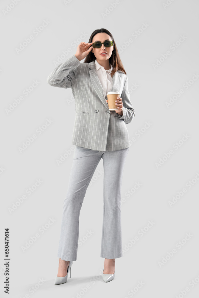 Young woman in stylish suit with cup of coffee on light background