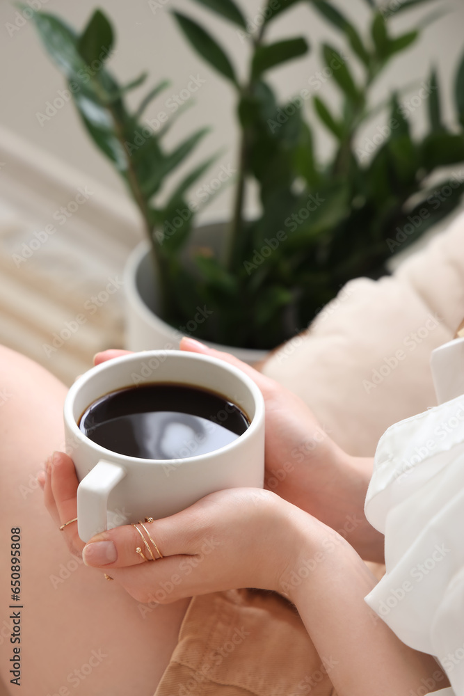 Young woman sitting in armchair and holding cup of delicious coffee