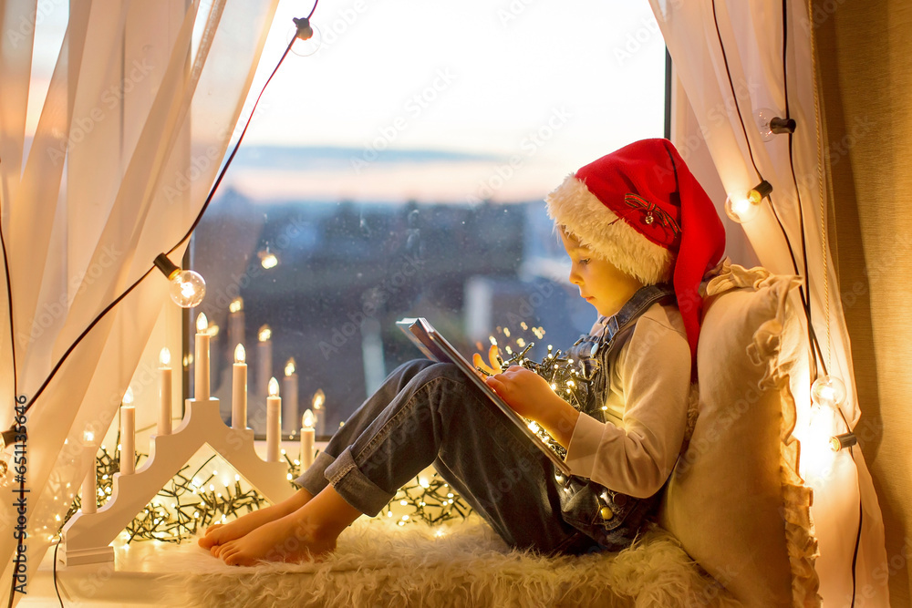 Toddler child, cute blond boy, sitting on the window, reading book, christmas lights around