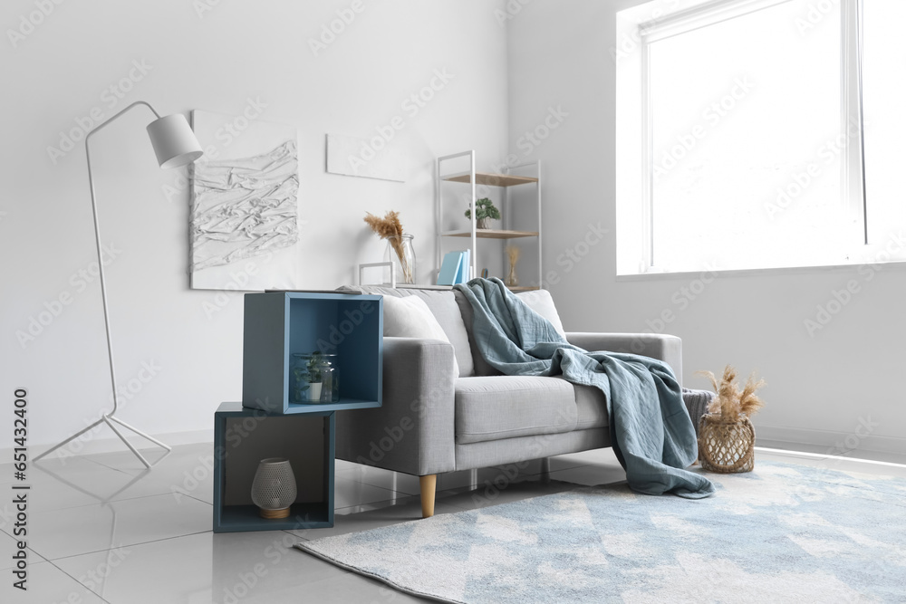 Cozy grey sofa with soft blanket, cushion and stylish shelves in interior of light living room