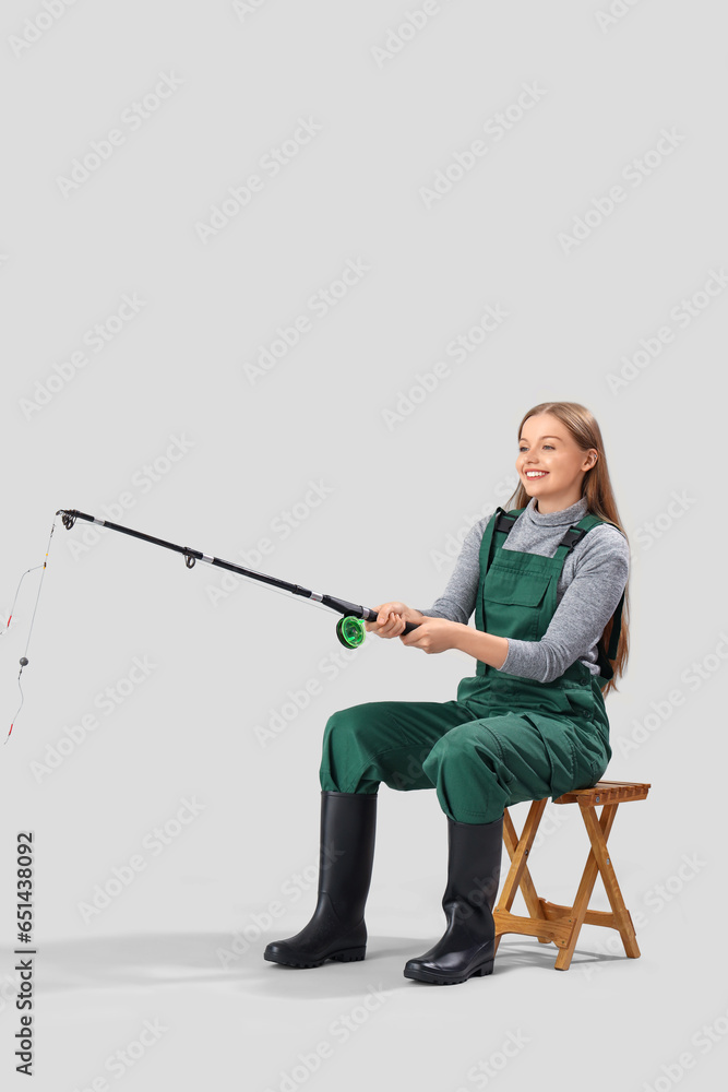 Young woman with fishing rod sitting on stool against light background