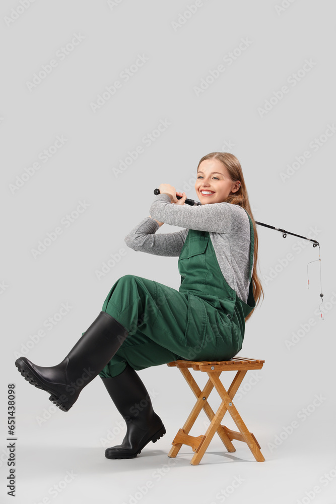 Young woman with fishing rod sitting on stool against light background