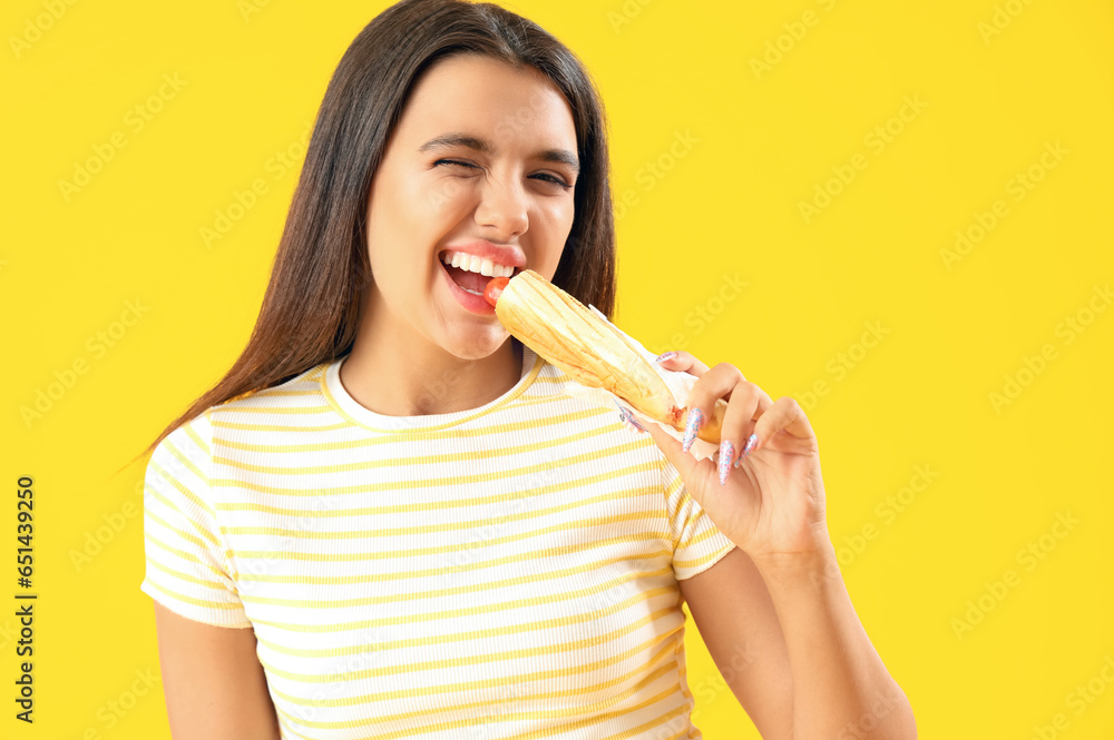 Pretty young woman eating tasty french hot dog on yellow background