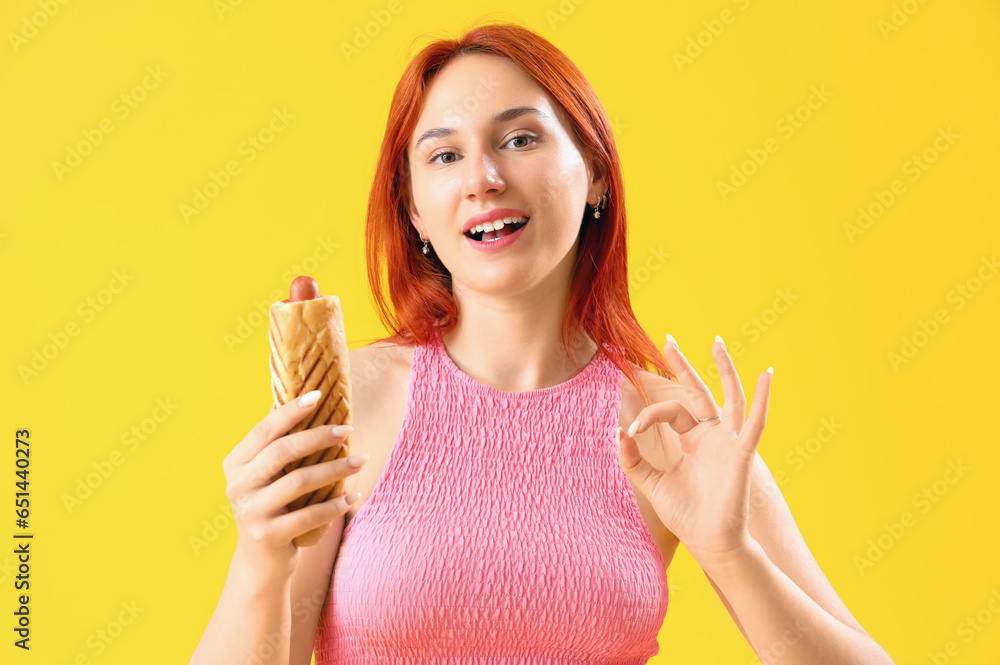 Beautiful young woman with tasty french hot dog showing OK on yellow background